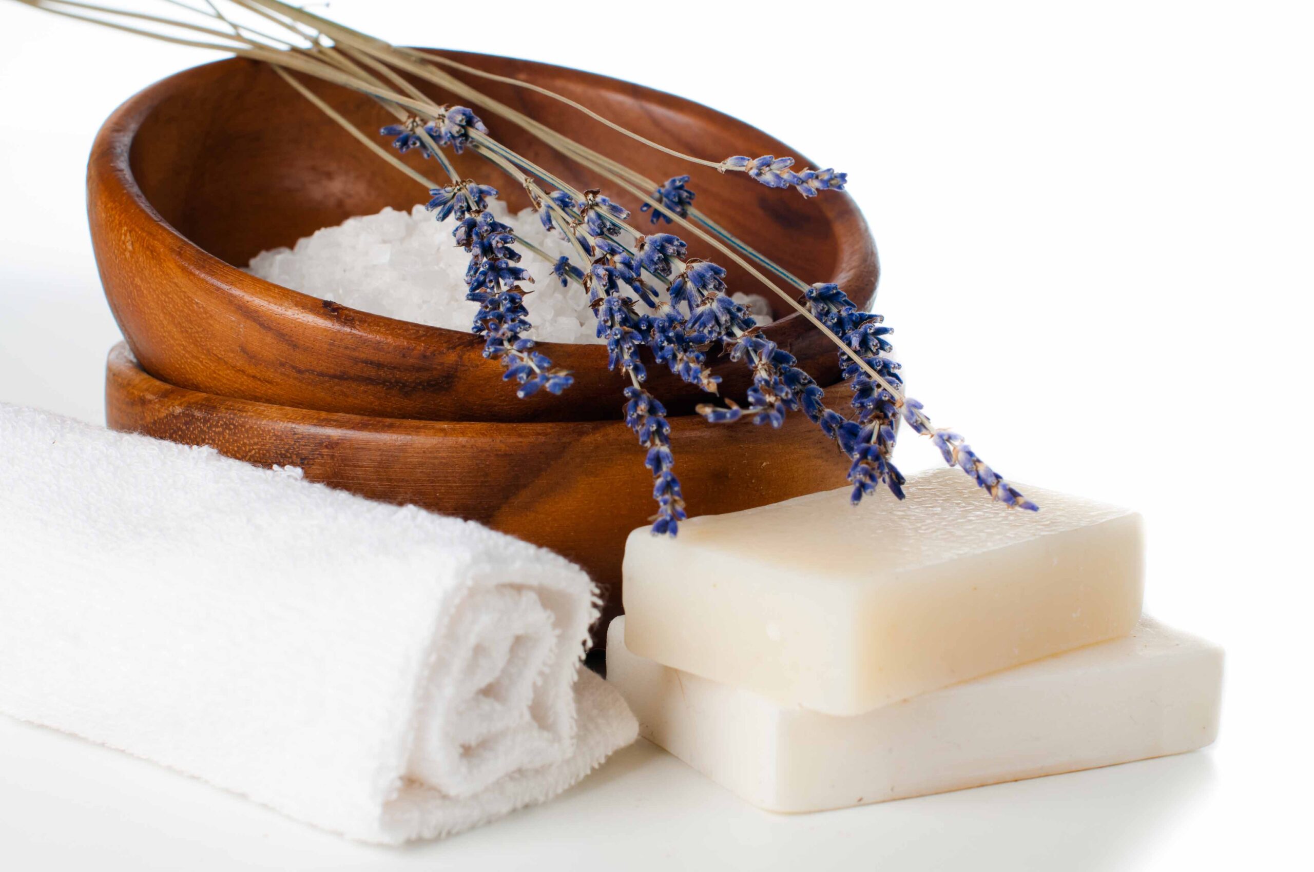 Magnesium bath salts and lavender in a wooden bowl next to a white bath towel and 2 new soap bars